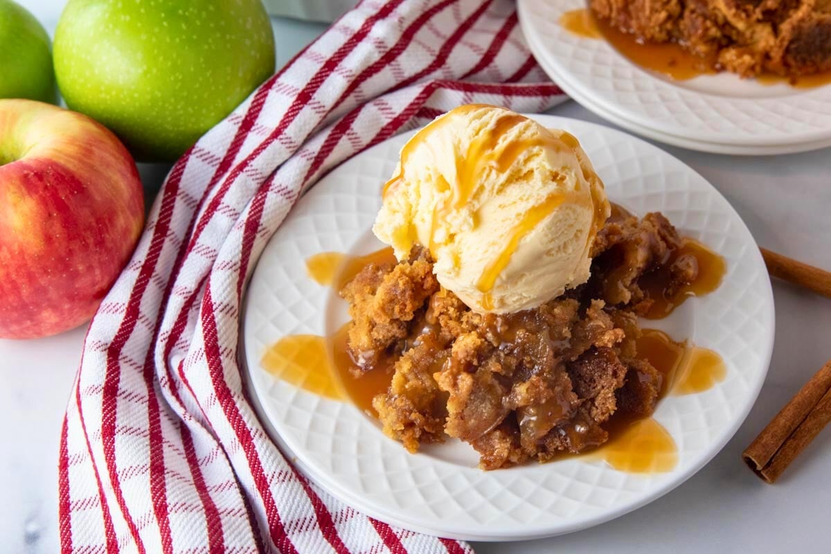 piece of caramel apple dump cake on a plate with ice cream and caramel sauce. Apples sitting beside.