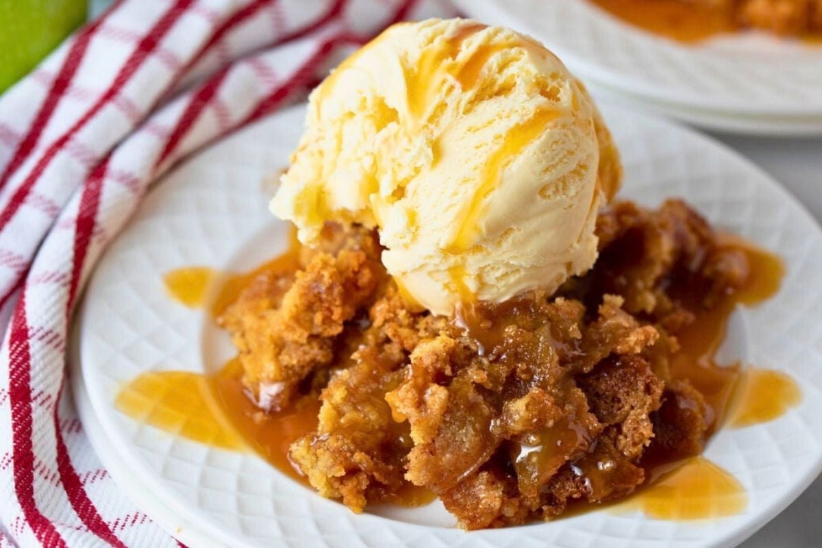 piece of caramel apple dump cake on a plate with ice cream and caramel sauce. Apples sitting beside.
