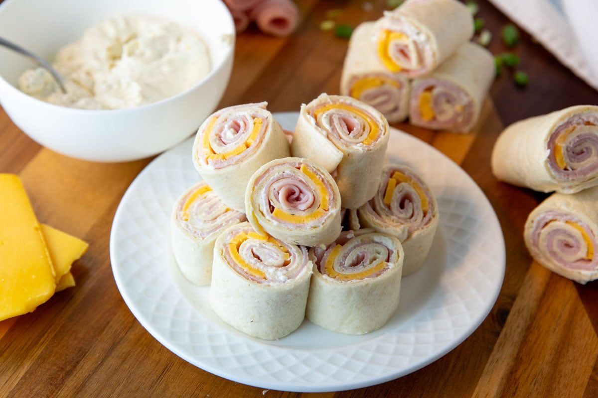 ham and cheese pinwheels on a plate with bowl of seasoned cream cheese, ham and cheese pinwheels, and slices of cheese surrounding.
