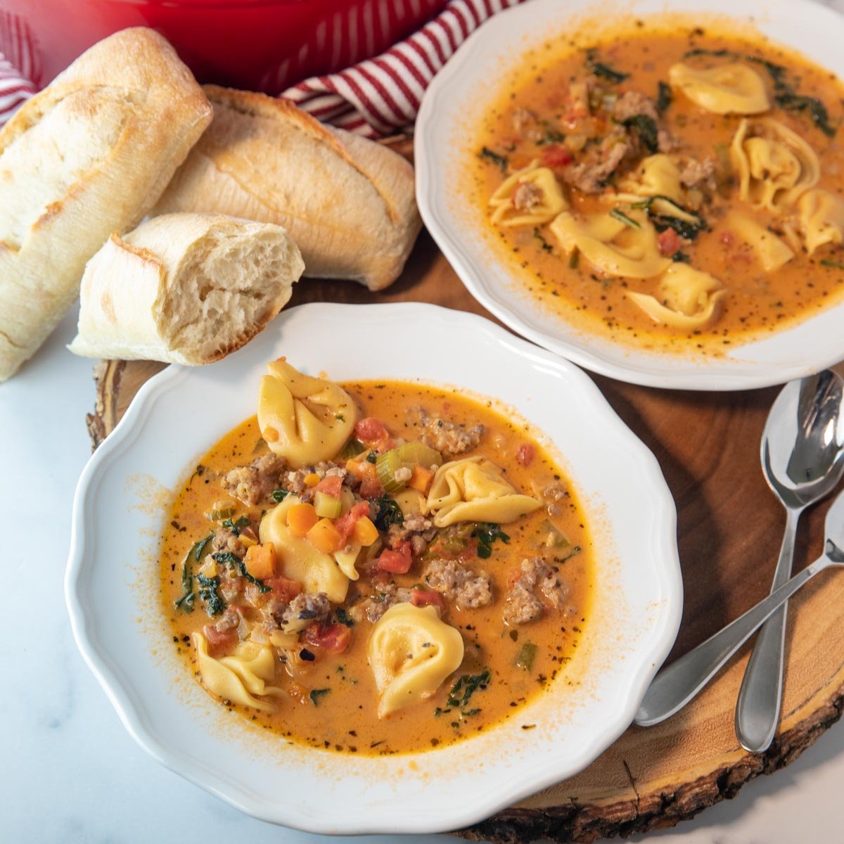 two bowls of spicy tortellini soup with bread and spoons sitting beside