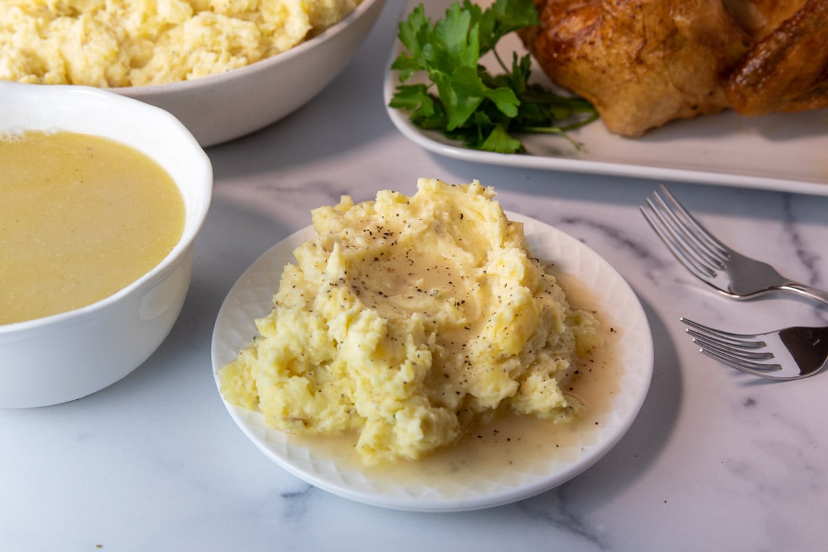 plate of mashed potatoes with turkey gravy. Bowl of turkey gravy and other Thanksgiving dishes sitting in background.
