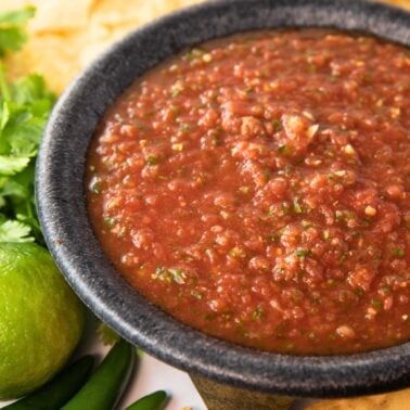 bowl of the best spicy salsa with chips, cilantro, lime, and peppers sitting beside.