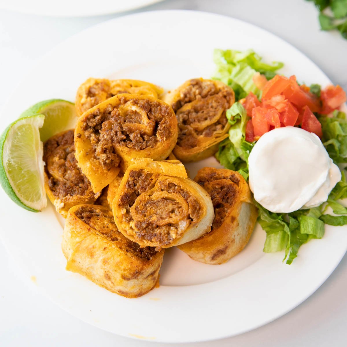 plate of taco pinwheels with lettuce, tomato, sour cream, and lime wedges sitting beside