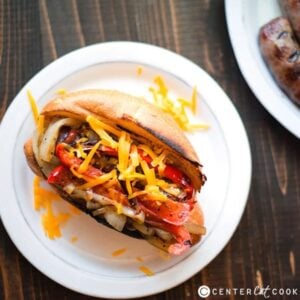 overhead of a bratwurst hot dog on a plate with sliced peppers and onions, ketchup, and mustard