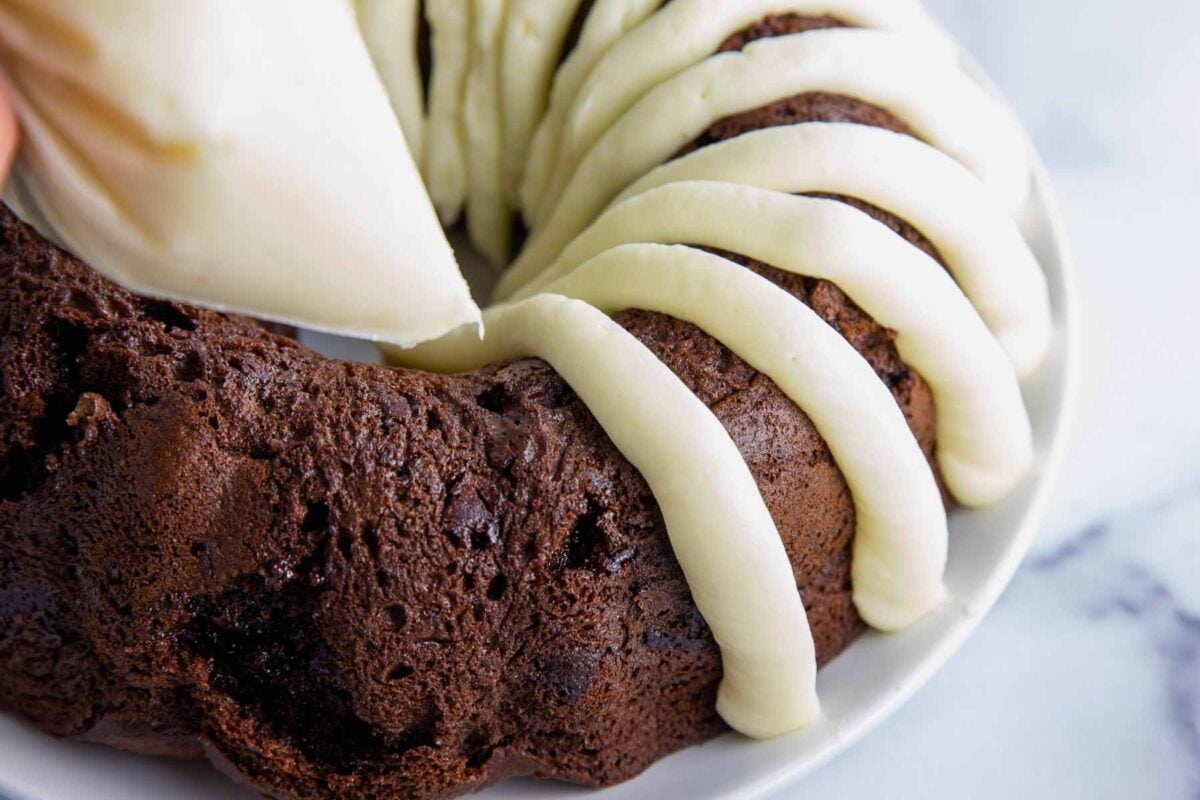 piping thick ribbons of cream cheese frosting onto a chocolate bunt cake in classic Nothing Bundt Cake Style.