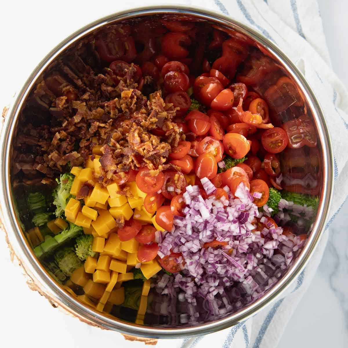 ingredients for broccoli salad in a mixing bowl including bacon, red onion, cheddar cheese and tomatoes.