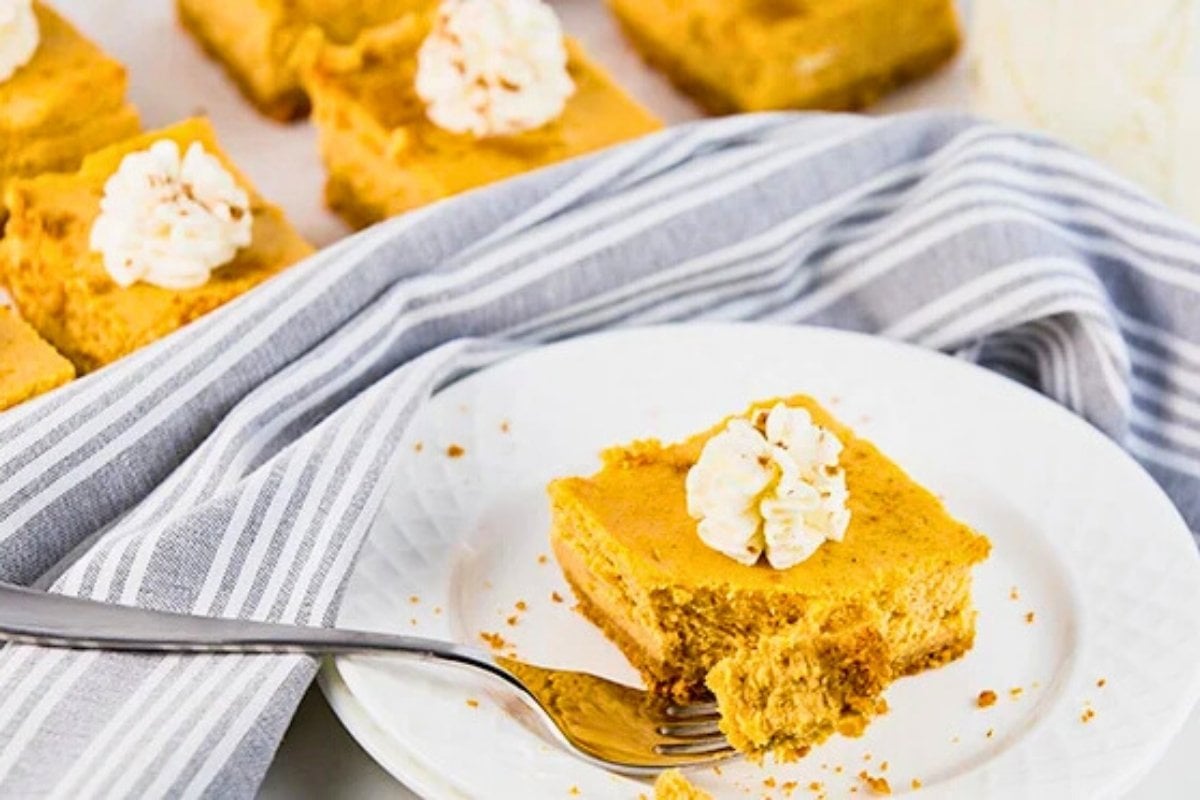 pumpkin cheesecake bars sliced into squares, served on a plate with whipped cream and pumpkin pie spice.